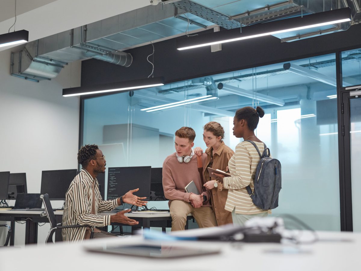 Wide angle view at diverse group of young people discussing project while working together in office or school IT lab with computers, copy space